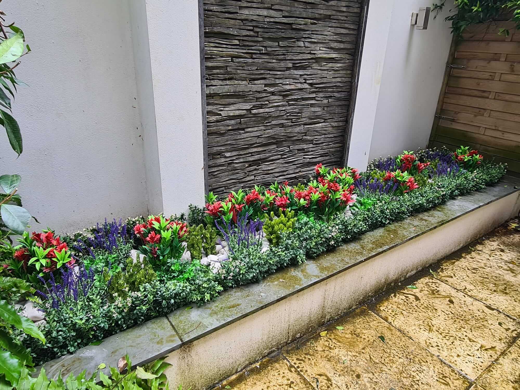 A rectangular flower bed with vibrant red and purple flowers, surrounded by lush greenery, is set against a white wall with a textured stone panel. The area is bordered by a stone path with a wooden fence to the right.