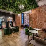 The Spring Pink Colour Box offers a stylish lounge with brick walls and wooden floors. Greenery adorns the ceiling, while plush seating complements marble tables. Cozy booths nestle by a patterned wall, with crystal chandeliers adding elegance to this chic sanctuary.