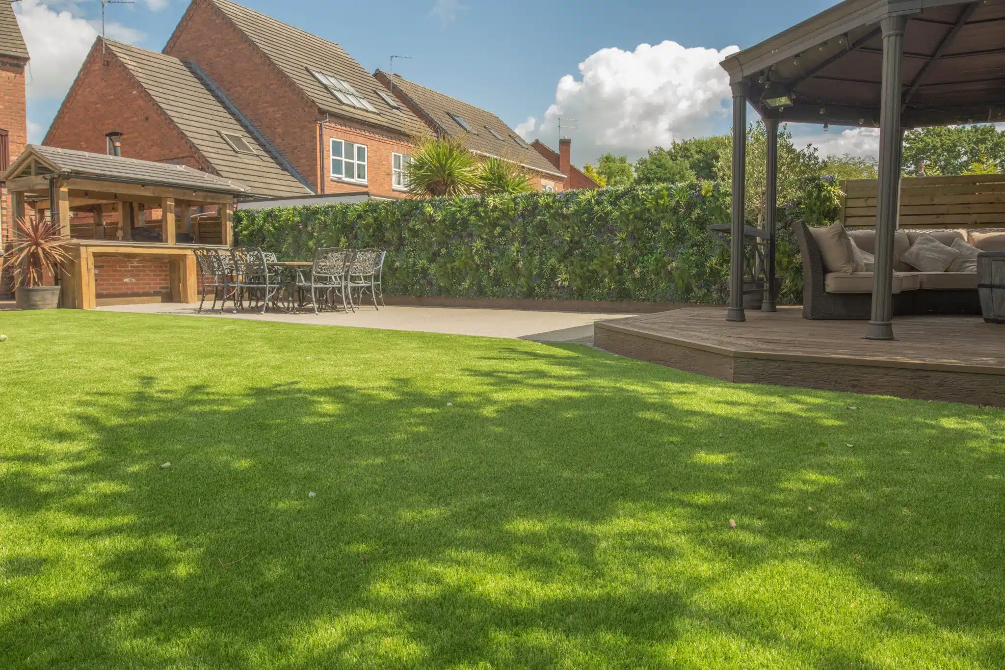 A backyard with a well-maintained lawn, a wooden gazebo to the right, and an outdoor dining area with chairs and a table to the left. Brick houses and a green hedge are in the background, under a partly cloudy sky.