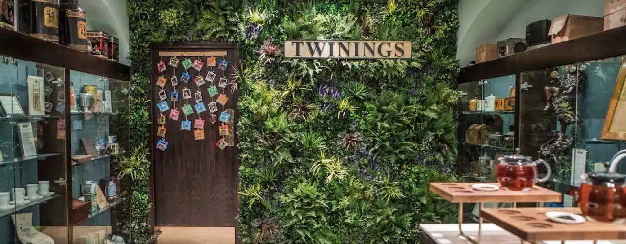 A room with lush green plants covering the walls and a sign that reads Twinings hints at how artificial plants are affecting store trends. Glass display cases on either side contain various items, while a wooden door at the end boasts colorful decorations.