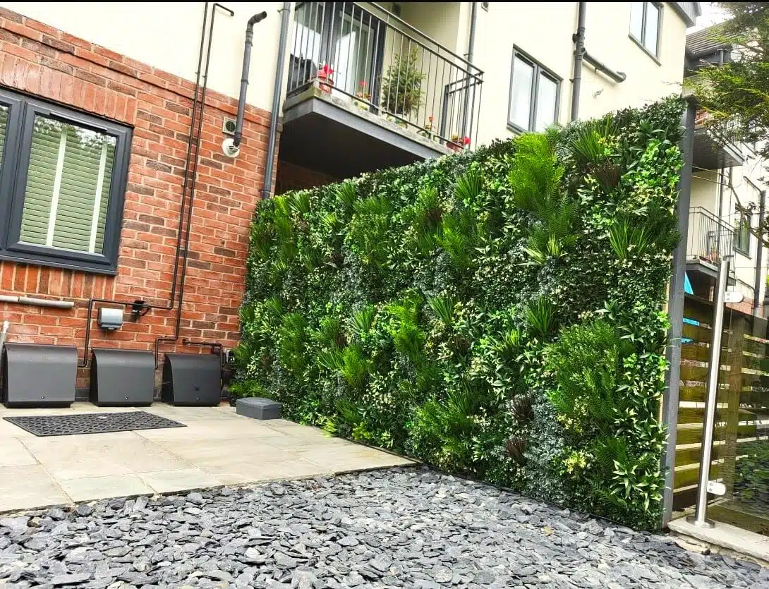 A vertical garden full of lush green plants adorns a wall in an urban courtyard. The space features a brick and cream-colored building, a small balcony with railings, and a stone patio with slate tiles and a few air vents.