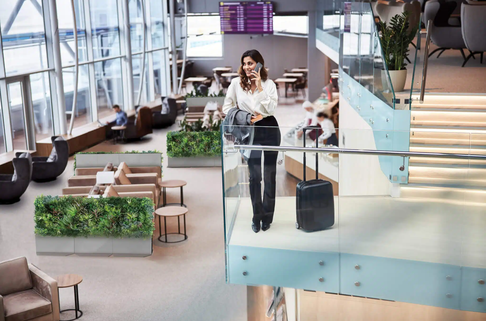 A woman in a white blouse and dark pants stands on an airport balcony, chatting on her phone. Next to her is a suitcase by Modular Green Wall Partitions, which add a natural touch to the scene. Background seats and a flight information board complete the setting.