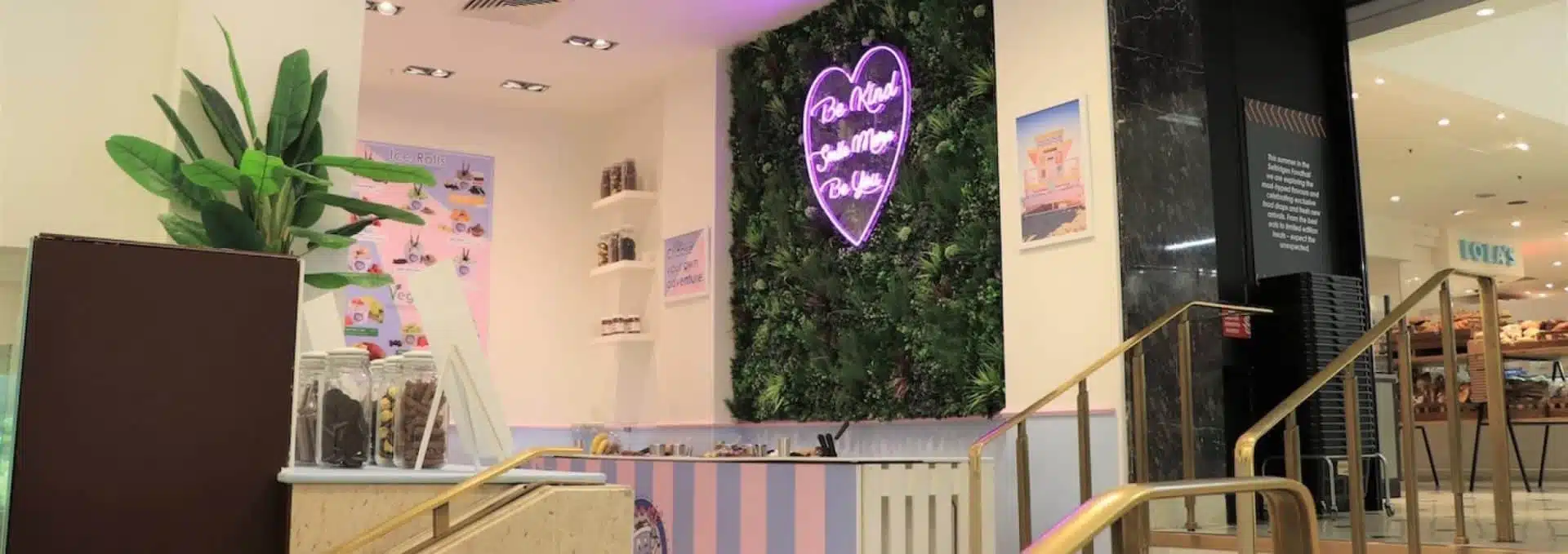 A modern retail store interior in London, with a neon pink heart sign reading Brooklyn Bamboo Baby on a lush green wall, perfect for social media. The space features striped pastel walls, a staircase with a gold railing, and various products displayed on shelves reminiscent of Selfridges style.