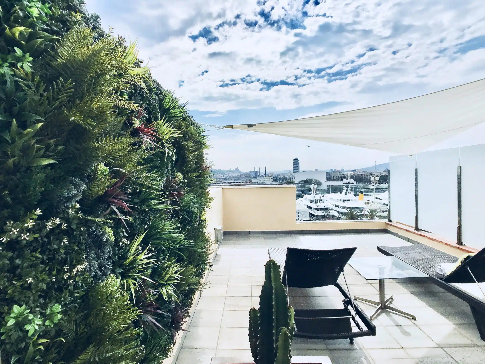 Rooftop terrace with a lush vertical garden wall on the left, overlooking a marina with boats. Two black lounge chairs and a small table are under a white shade sail. The sky is partly cloudy.