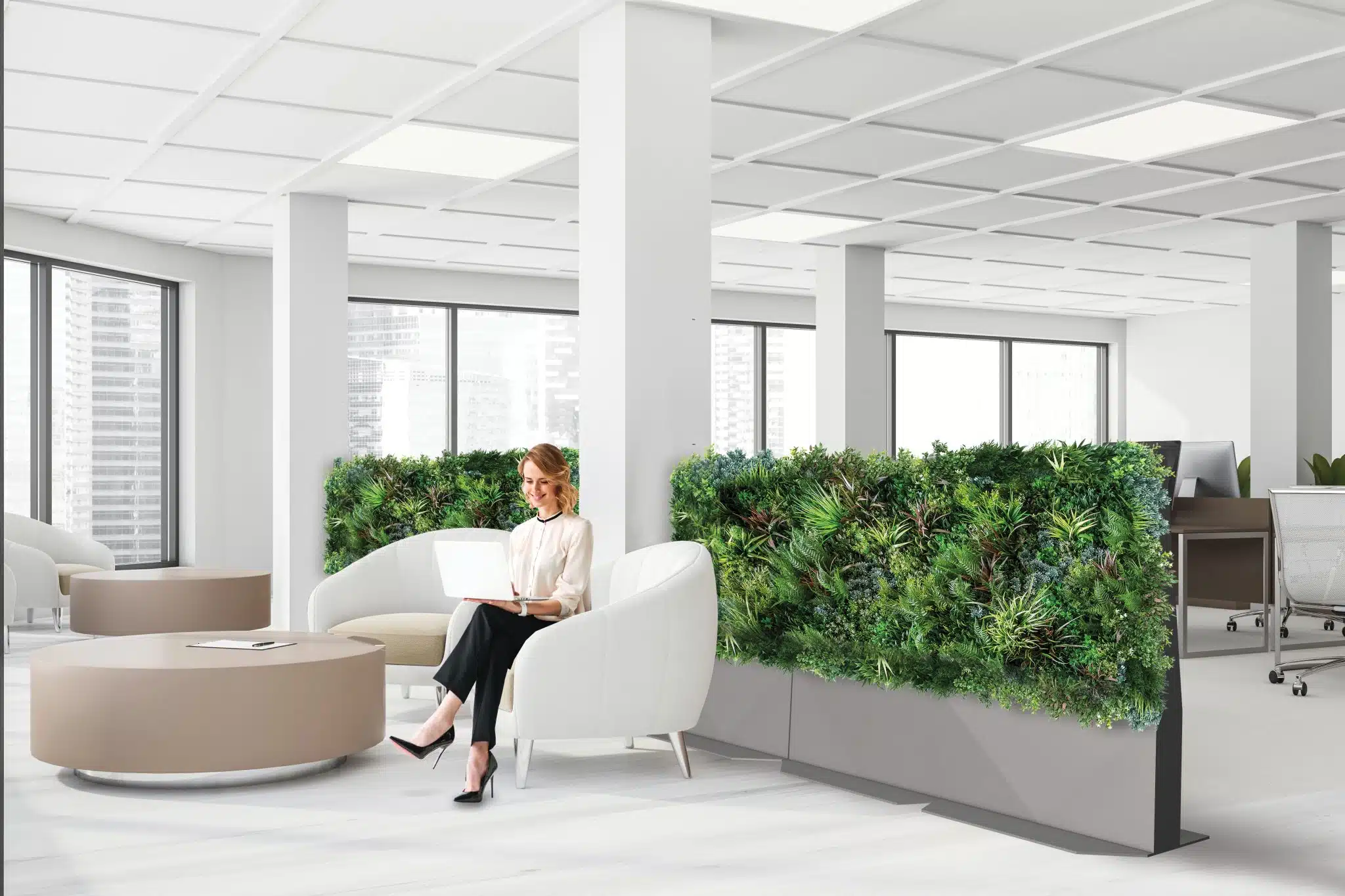 Seated on a white chair in a bright office, a woman uses her laptop near lush Modular Green Wall Partitions. Nearby are round tables and additional seating, with large windows that provide stunning cityscape views.