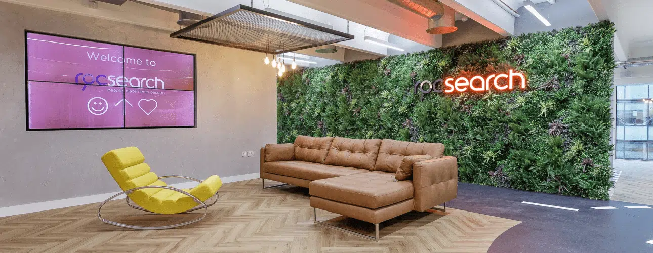 In a modern office reception, the space boasts a tan leather couch and yellow rocking chair against a vibrant vertical garden wall. The Vistafolia panels elevate acoustic qualities, ensuring tranquility. A pink screen warmly displays 