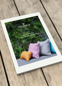 A brochure on a wooden table showcases a cover with an artificial green plant wall and colorful cushions. The text reads: Vistafolia: Practical Greening to Create Amazing Environments Inside & Out.