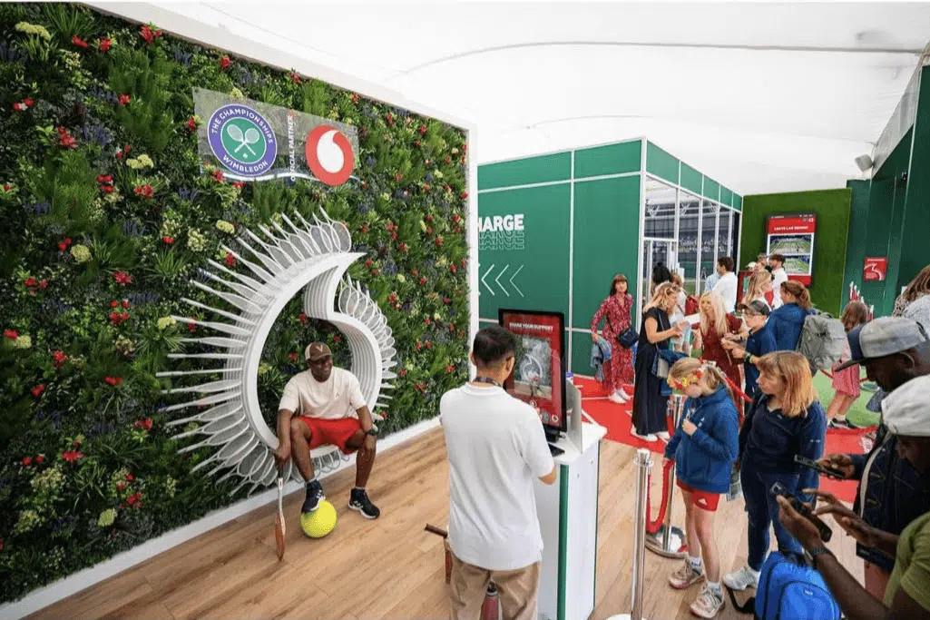 A man sits on a chair shaped like the Vodafone logo in front of a lush, green wall featuring Wimbledon and Vodafone logos. People wait in line, interacting with digital screens. The venue has a modern, open booth design.