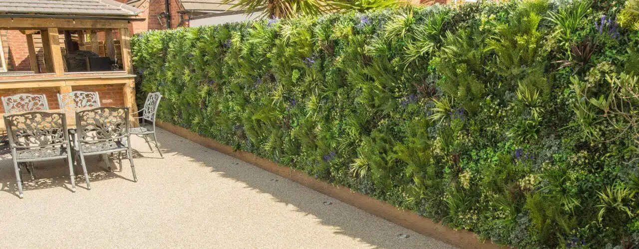 A garden patio features metal chairs and a decorative metal table, while a lush vertical garden wall brimming with diverse plants lines one side. This vibrant natural backdrop, under a sunny sky, illustrates the top benefits of vertical gardens in enhancing small outdoor spaces.