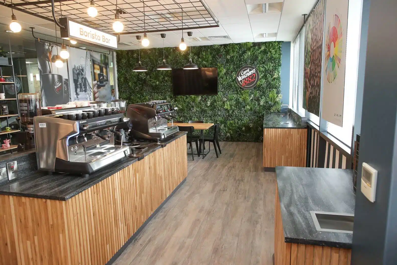 A modern café with wooden countertops and espresso machines features lush green plant walls, reflecting the Café Green Walls in Ireland vibe. A Lavazza logo adorns the wall above a TV. Bright lights illuminate the wooden floors and a cozy seating area at the back, offering coffee with a view.
