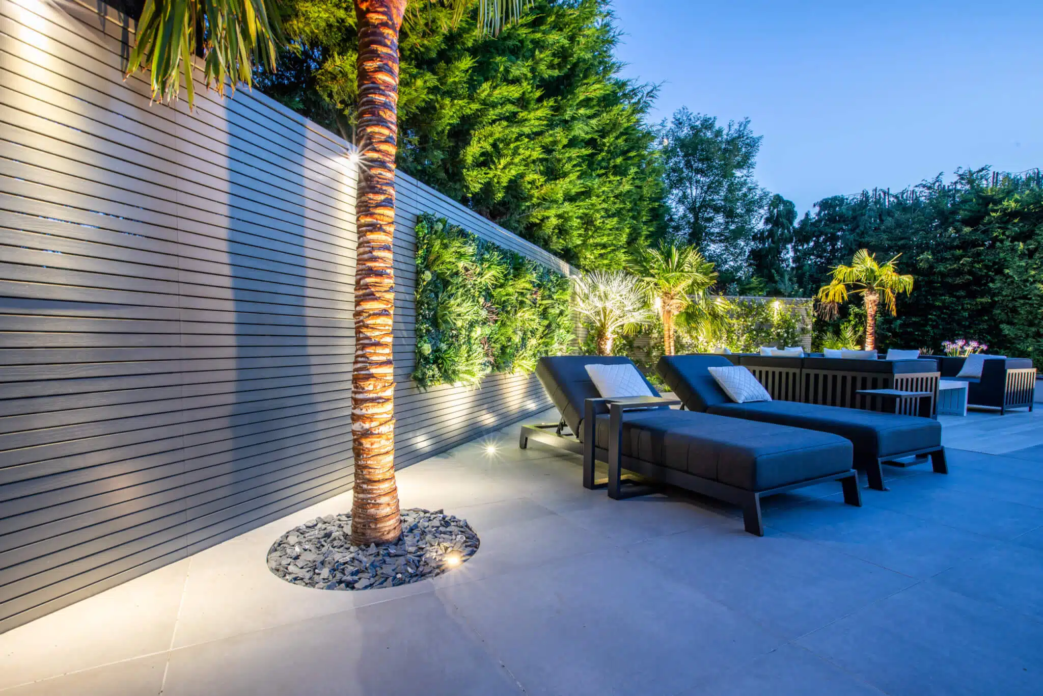 Modern outdoor patio with two dark lounge chairs on a tiled surface. A lit, striped palm tree stands nearby, surrounded by greenery. Ambient lighting and a wooden slat fence enhance the tranquil setting.