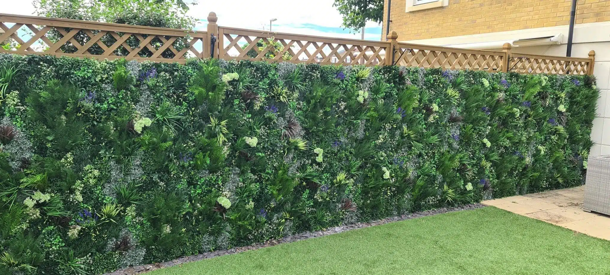 A garden with a tall vertical fence blossoming in dense, green foliage and vibrant flowers, thanks to an expert artificial green wall installation. The ground is lush with grass, complemented by a charming wooden lattice fence crowning the verdant wall.