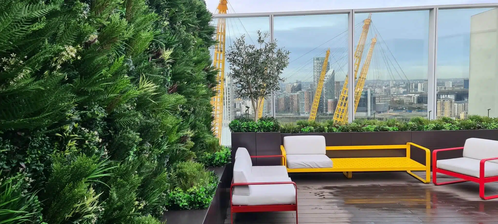 Greenwich rooftop patio with an artificial green wall on the left, offering a stunning view of a city skyline and cranes in the background. The City Roof Terrace boasts colorful seating with red, white, and yellow cushions on a wooden deck.