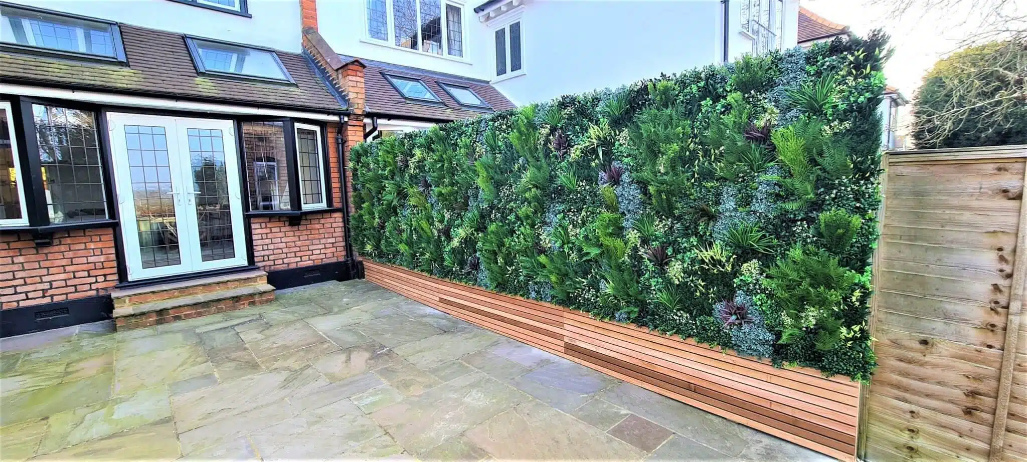 A patio area with stone paving next to a building with white-framed doors and windows. A lush green artificial green wall with various foliage types sits above a wooden panel, creating a vibrant garden feature beautifully complemented by sleek fencing.