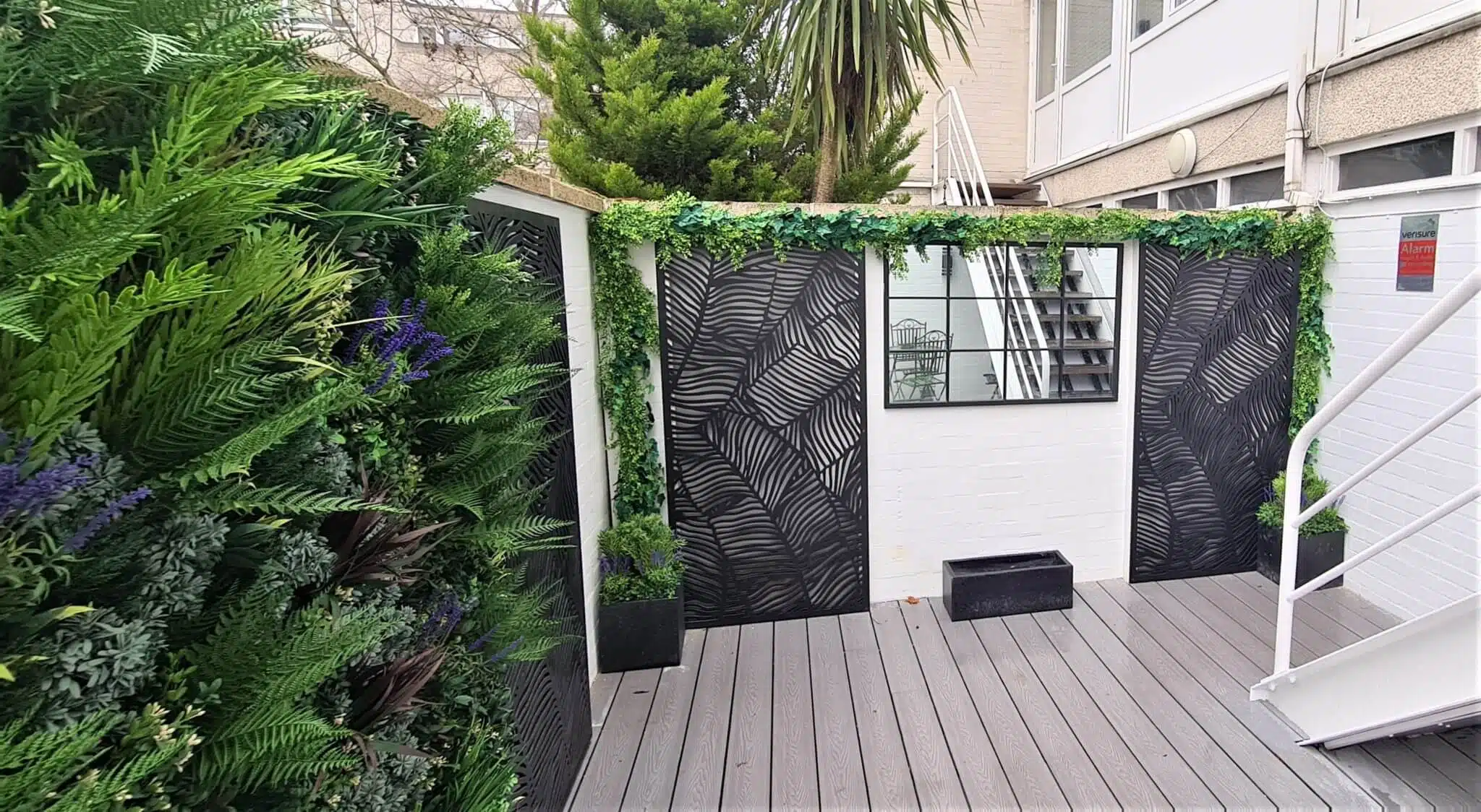 A low-maintenance Windsor courtyard garden, this small outdoor patio features gray decking surrounded by black metal panels with leaf patterns. Greenery and purple flowers adorn the left wall, while plants grace the top. A mirror decorates a white brick wall at the back, and white stairs sit on the right.