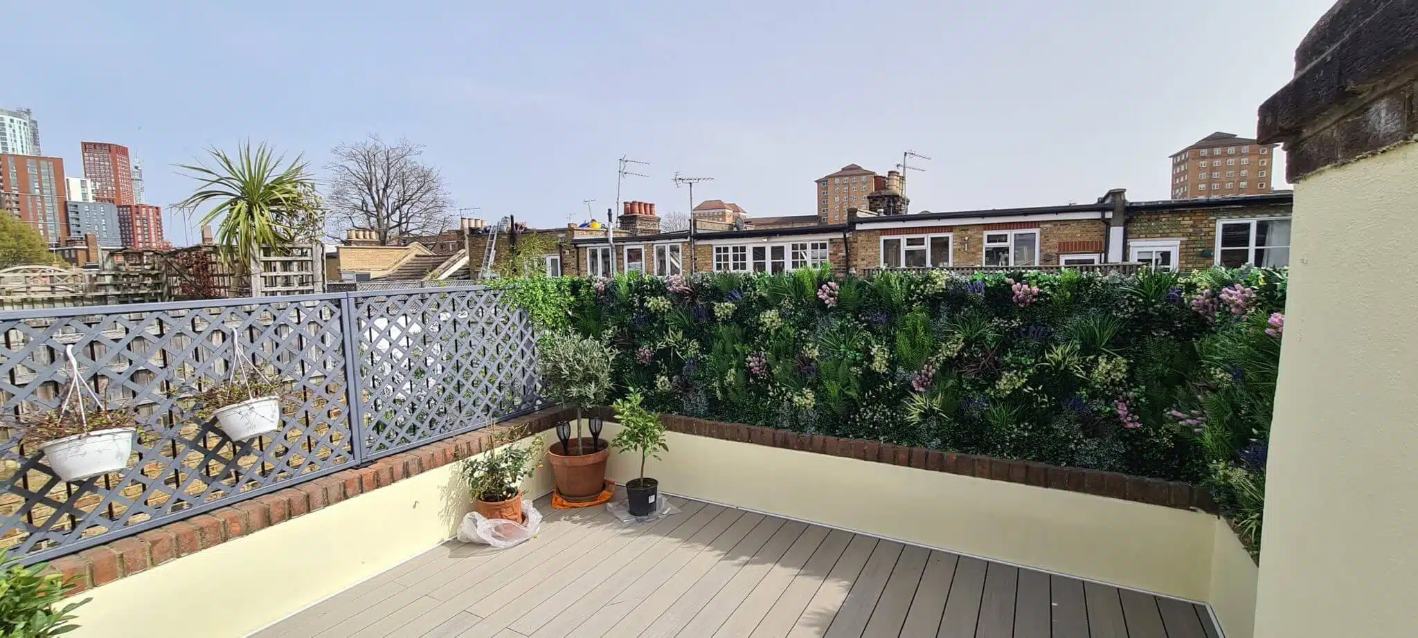 Rooftop patio with wood-like flooring, enveloped by a decorative lattice fence on the left and a vibrant green wall on the right. Potted plants adorn the corners. The backdrop showcases urban London buildings under a clear sky, epitomizing an idyllic roof terrace retreat.