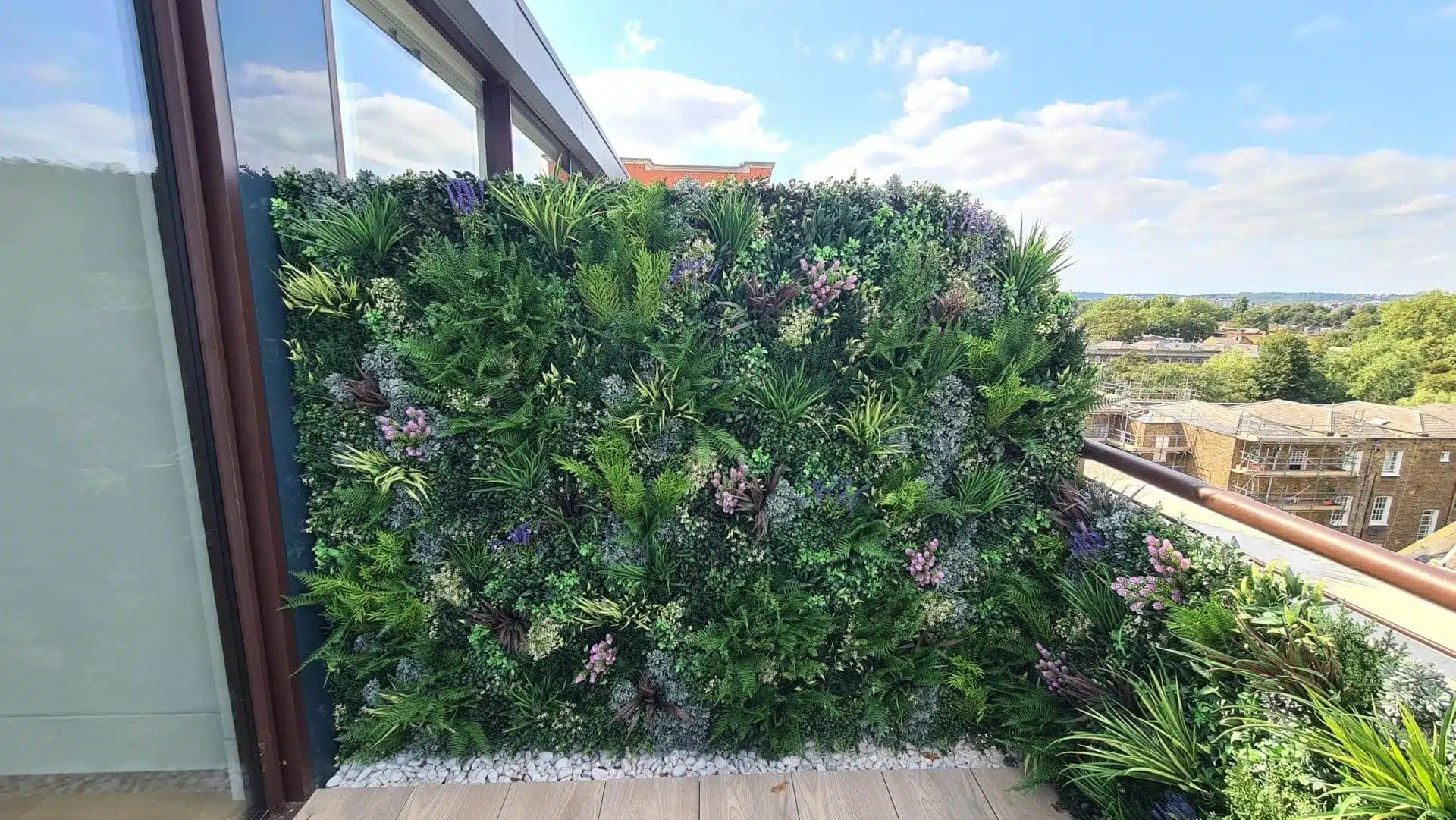 A vibrant green wall installation, adorned with ferns and colorful flowers, graces a London balcony. It overlooks the cityscape, where historic buildings meet patches of greenery under a blue sky dotted with scattered clouds.