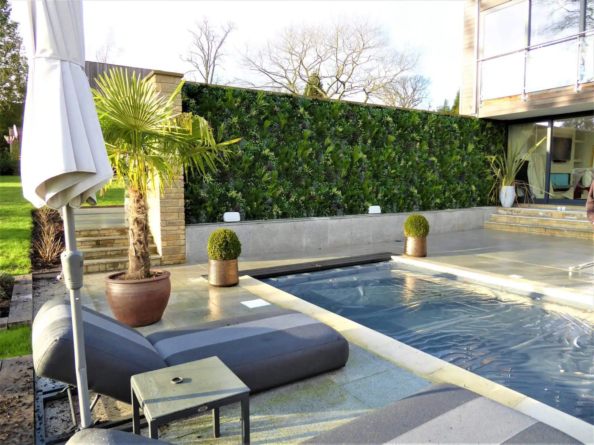 Modern outdoor patio with a small pool, complemented by an artificial green wall installation. Lounge chairs, a potted palm, a white umbrella, and a glass railing add to the ambiance. The scene is bright with clear skies and tall trees in the background.