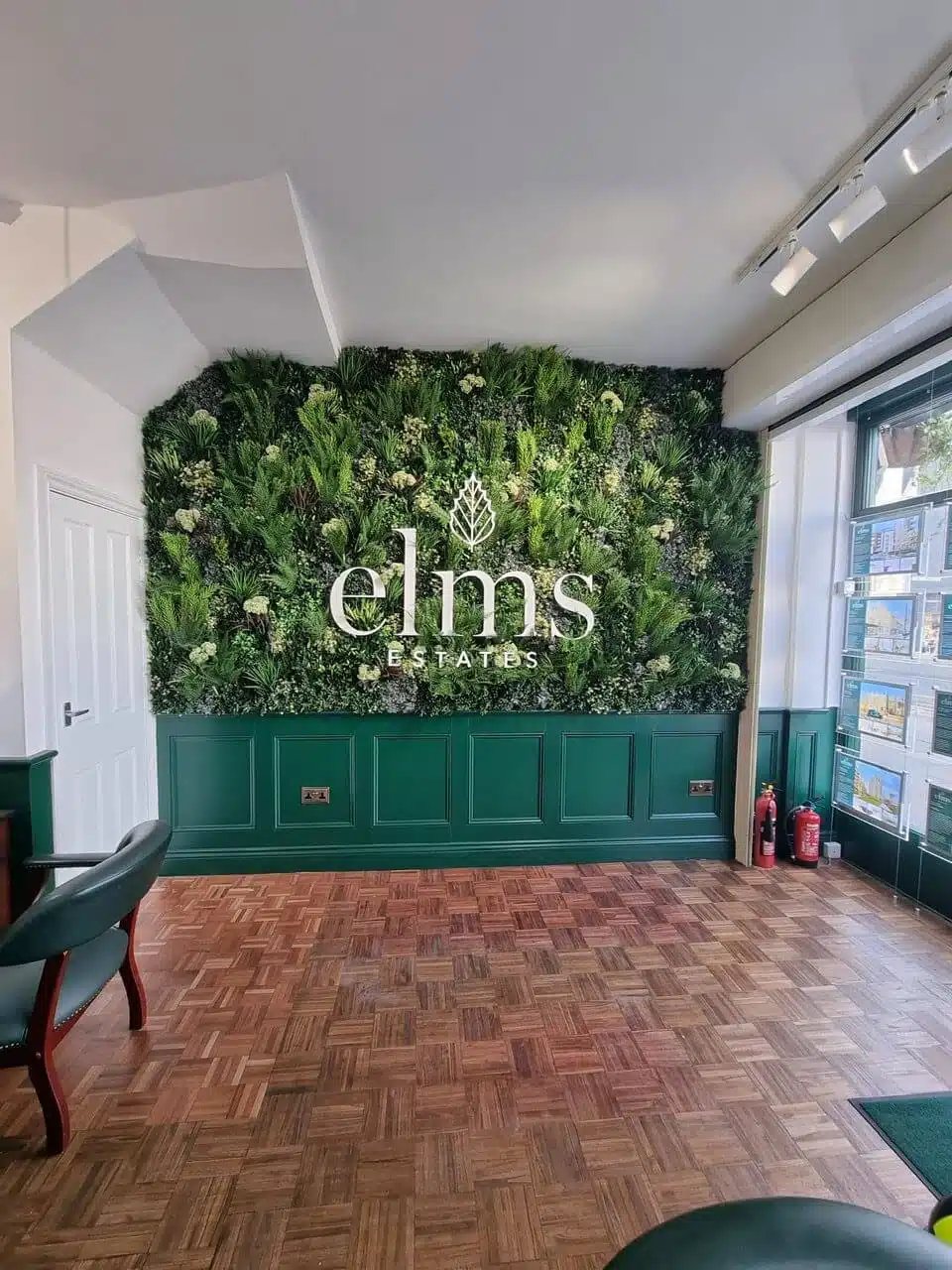 A room features a green wall with signage proclaiming elms ESTATES, capturing the essence of a London estate agents office. The wooden floor contrasts with two red chairs, while fire extinguishers stand in the corner. A large window on the right offers an inviting view outside.