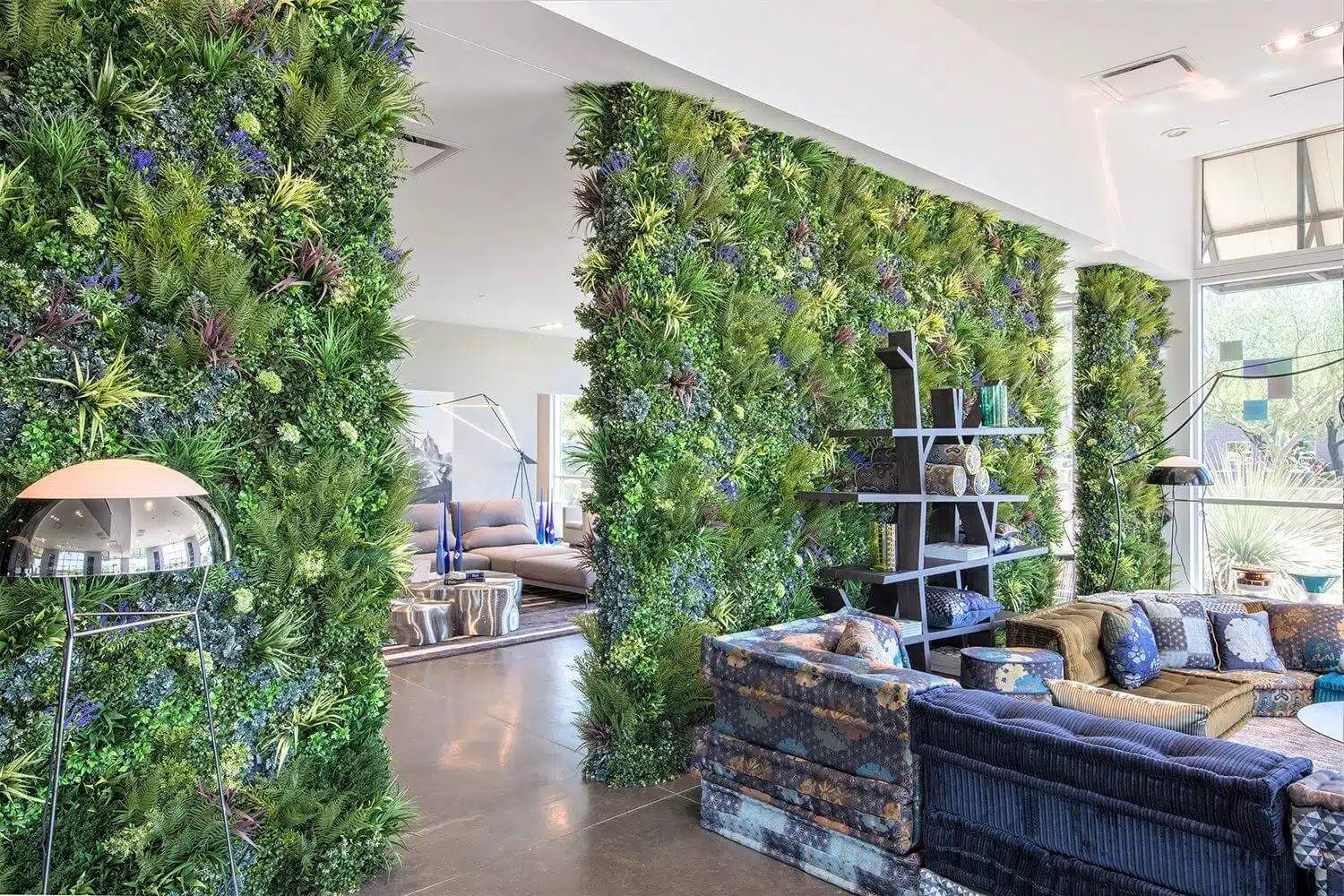 A modern living space with green plant walls separates two areas. On one side, a seating area with blue-patterned sofas and shelves; on the other, a cozy living area with a floor lamp and gray couches. Large windows bring in natural light.