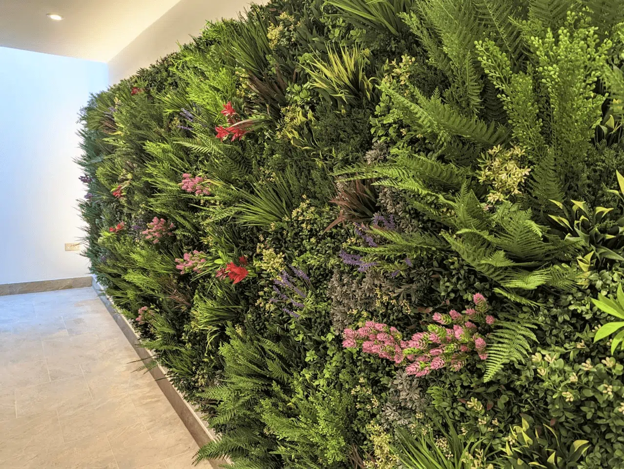 A lush residential faux living wall in Kent, featuring a variety of green ferns, leafy plants, and vibrant flowers in red, pink, and purple hues. The garden is set against a neutral-toned wall with a light-colored tiled floor.