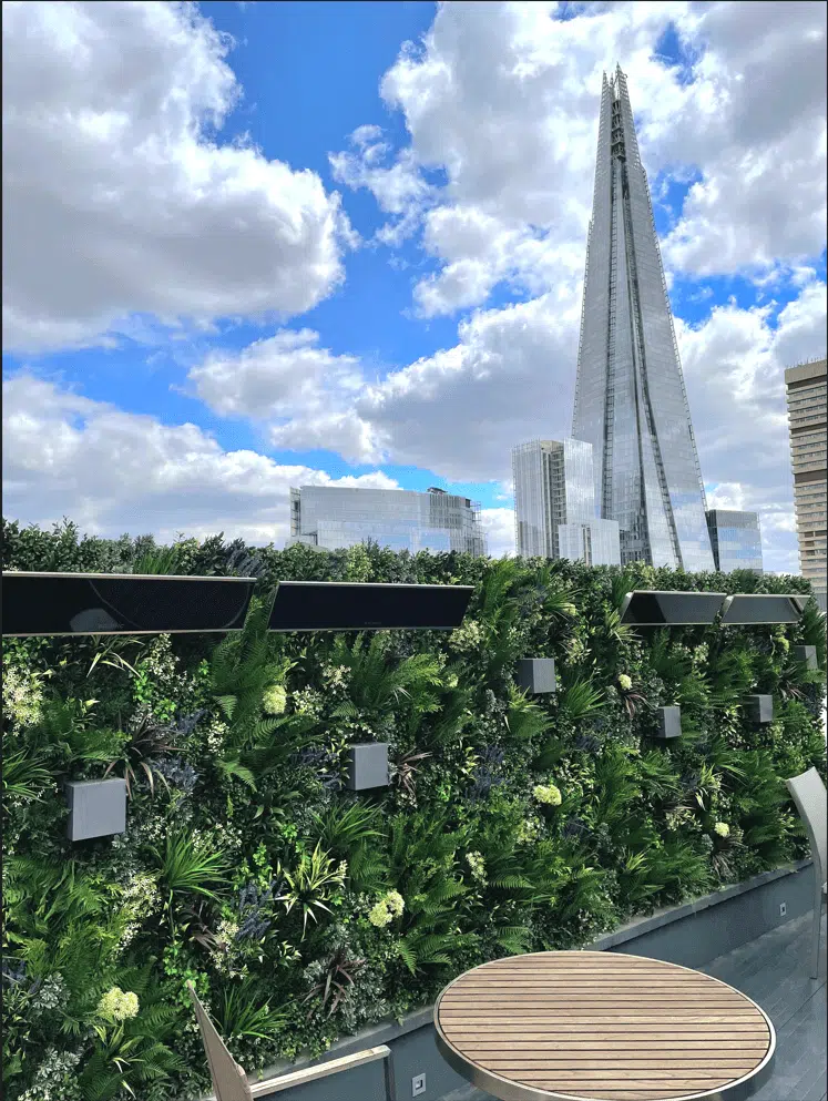 On a sunny day with scattered clouds, the skyline image showcases The Shard skyscraper in London. In the foreground, a lush green wall and round wooden table highlight a charming residential roof terrace in Southwark, contrasting beautifully with the modern cityscape.