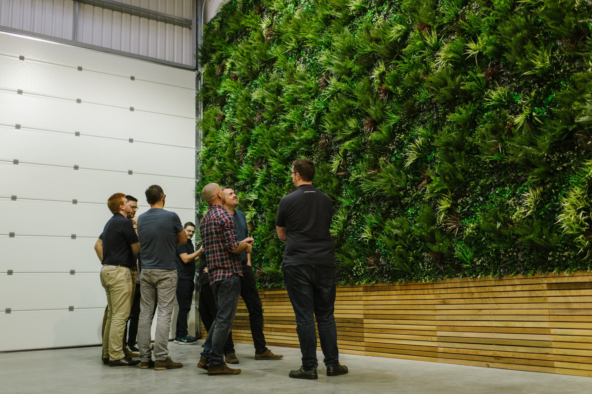 A group of people stands in a modern space with concrete floors, observing a large green wall of lush plants. Wooden paneling borders the base of the vertical garden.