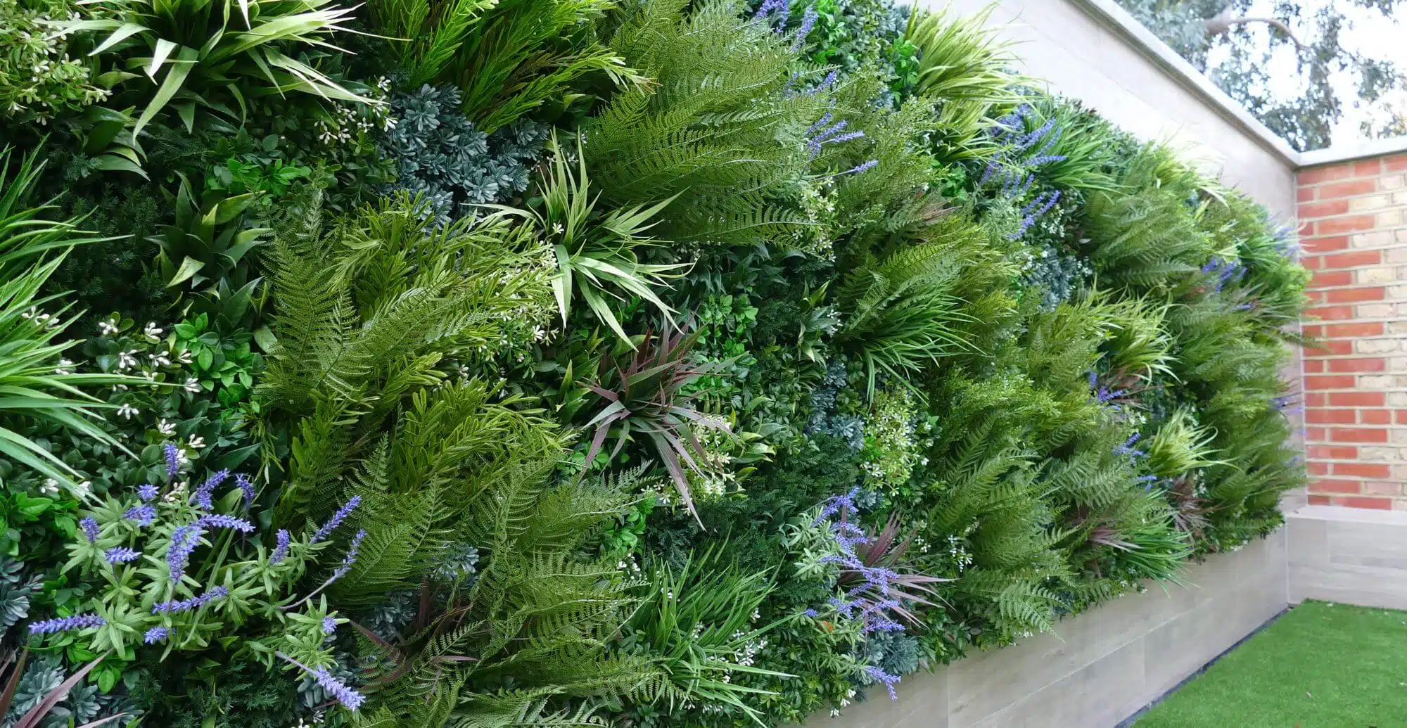 A lush vertical garden covers a wall with a variety of green plants and clusters of small purple flowers. A brick wall and a patch of green grass are visible in the background, creating a vibrant outdoor setting.