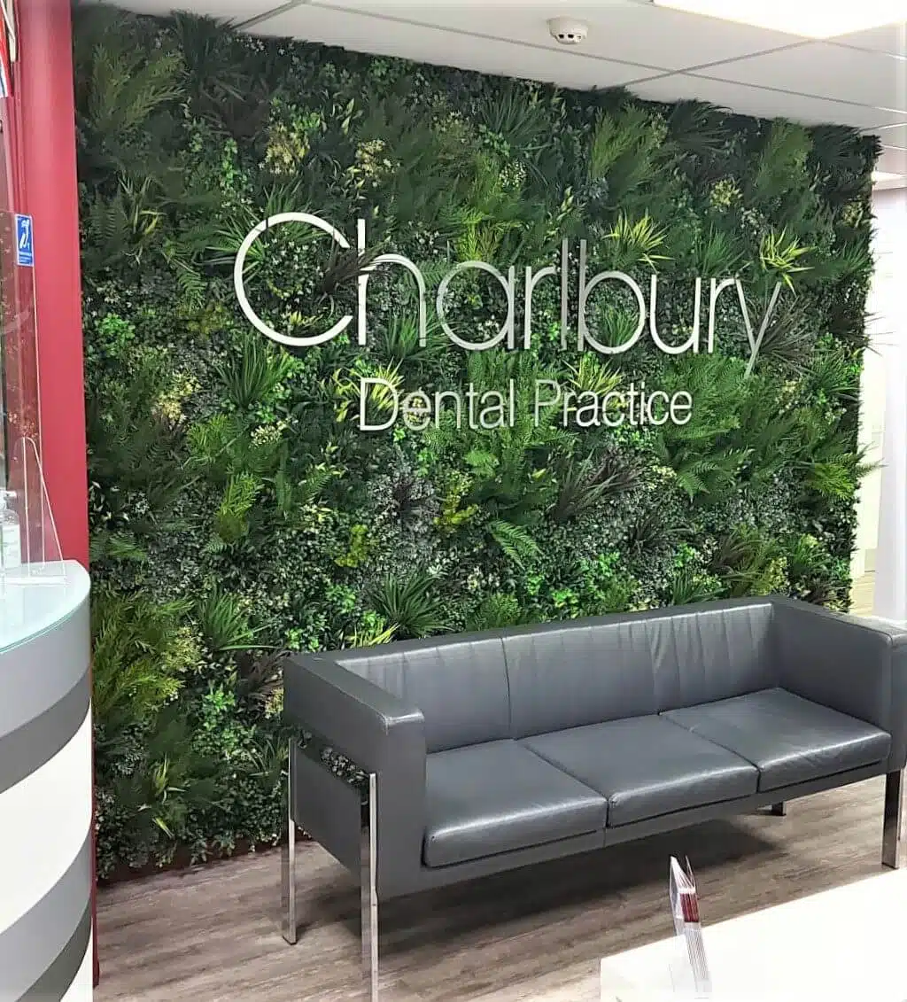 A modern dental office waiting area in Oxfordshire features a gray sofa and a lush green plant wall showcasing Charlbury Dental Practice. The interior boasts a minimalist design with sleek wood flooring.