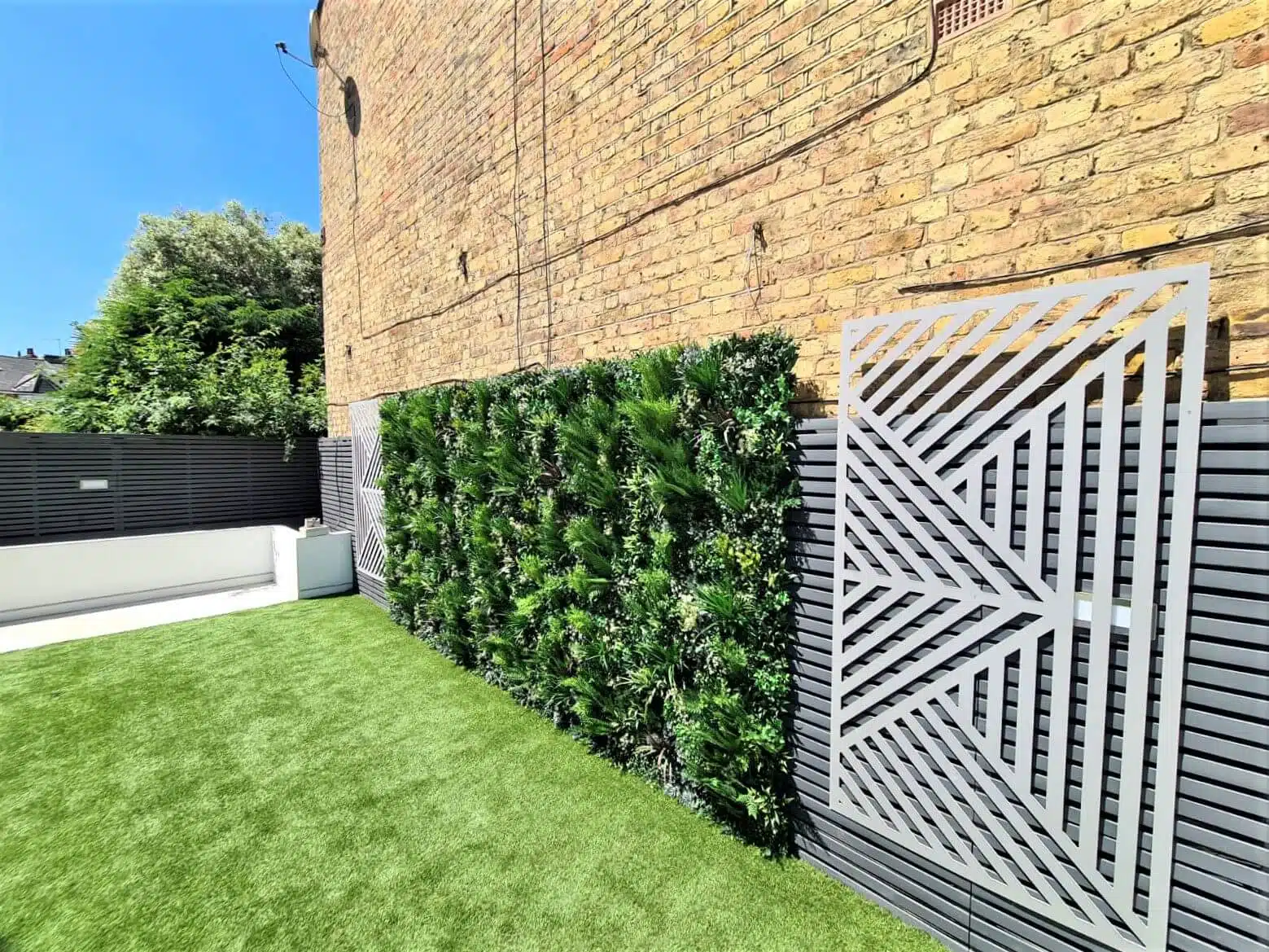 A garden in Hampstead with artificial grass, a planted green wall, and a decorative metal panel fence. The backdrop showcases a tall brick wall and the endless charm of a clear blue sky above.