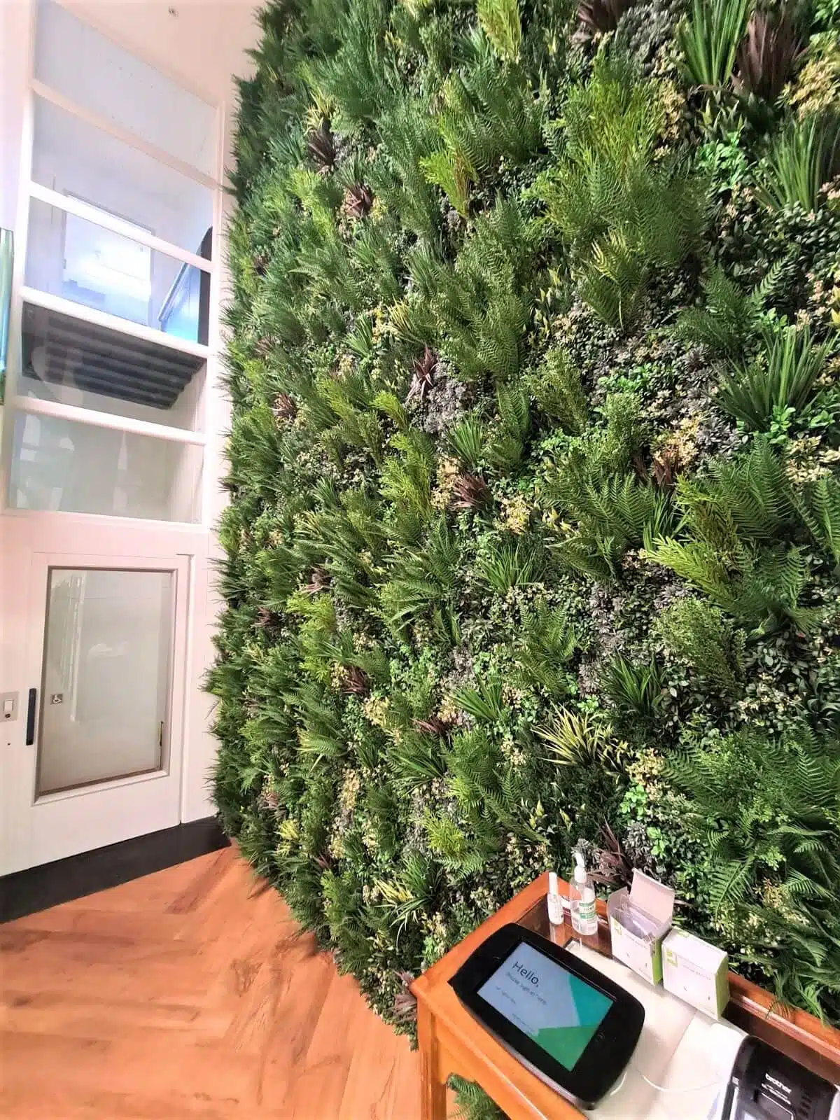 At The RSBC Headquarters in London, a large indoor vertical garden flourishes with lush green plants covering a wall beside a wooden floor. A small wooden table holds a tablet with information, surrounded by boxes and bottles. A white shelving unit stands to the left.