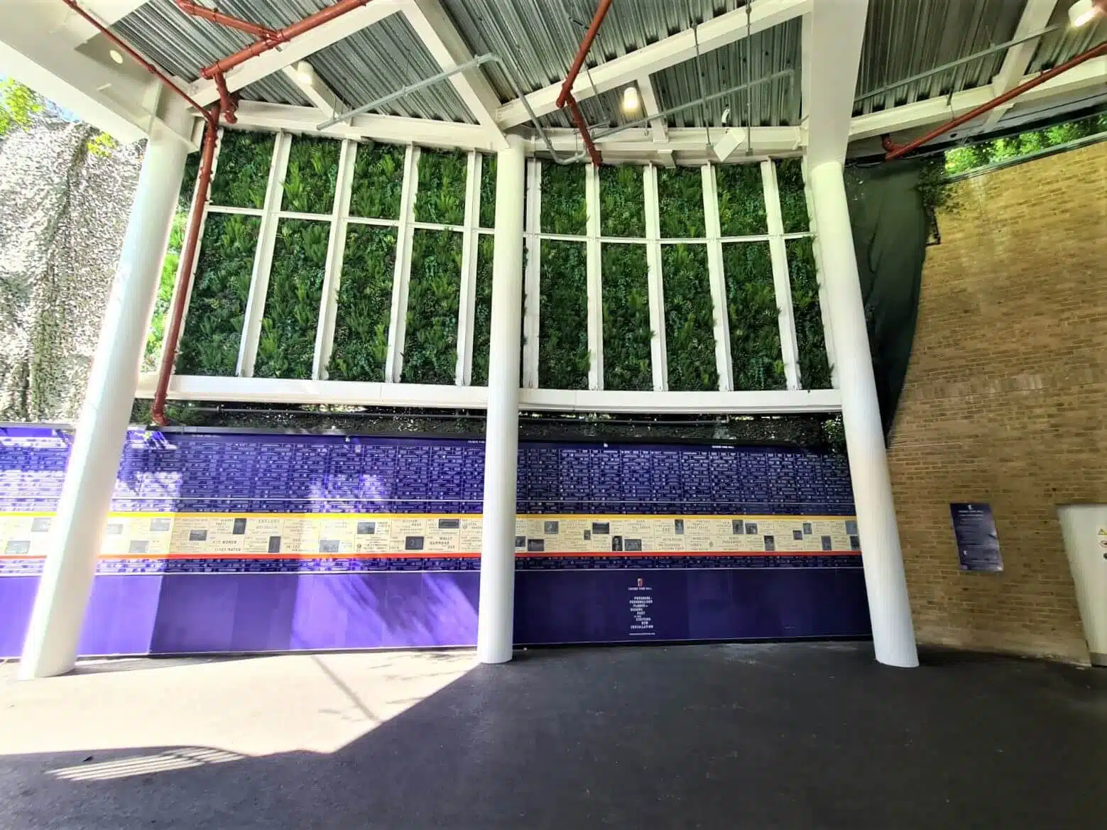A covered outdoor space with a living green wall supported by white columns at what feels like Londons historic Lords Cricket Ground. Behind the columns, a mural showcases blue tiles with yellow accents and historical plaques. The floor is paved, and a brick wall is partially visible.