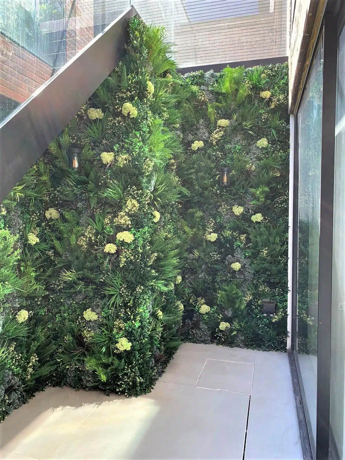 A basement green wall installation in Wimbledon showcases an indoor vertical garden with lush green foliage and white flowers covering a tall, corner wall. Sunlight streams through the glass ceiling and walls, illuminating the space as a narrow concrete floor leads to the corner.