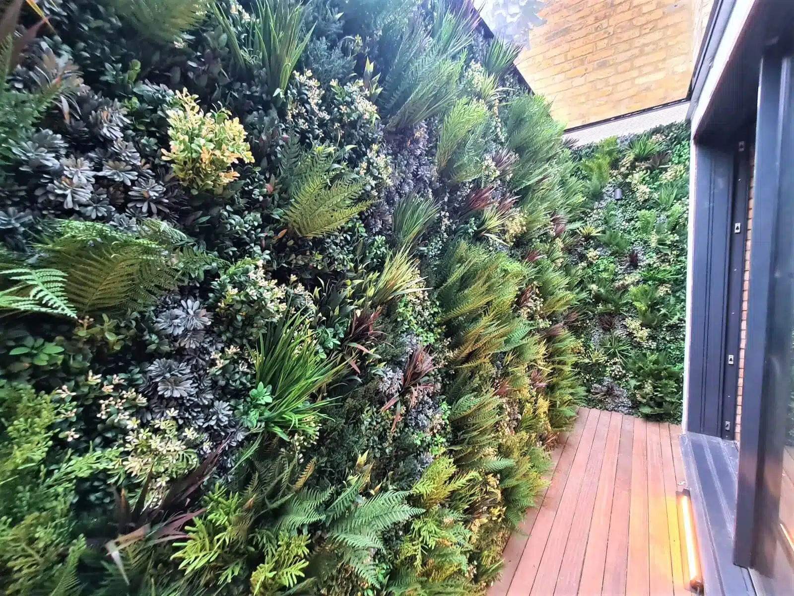 A narrow corridor in a London lightwell features vertical walls covered in lush green plants, including ferns and small shrubs. The wooden floor contrasts with the greenery, while a glass skylight allows natural light to illuminate this green wall installation.