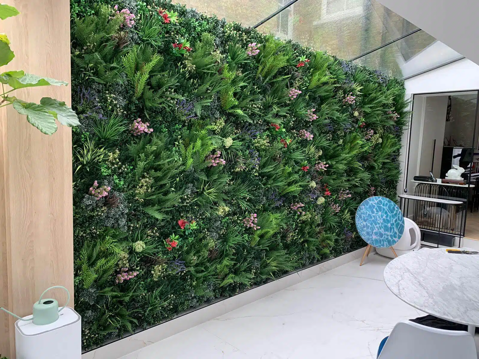 A lush vertical green wall with vibrant plants and pink flowers graces a bright indoor conservatory. A white round table sits in the foreground, complemented by a potted plant and a uniquely designed chair with a blue seat, exemplifying Fulhams chic aesthetic.