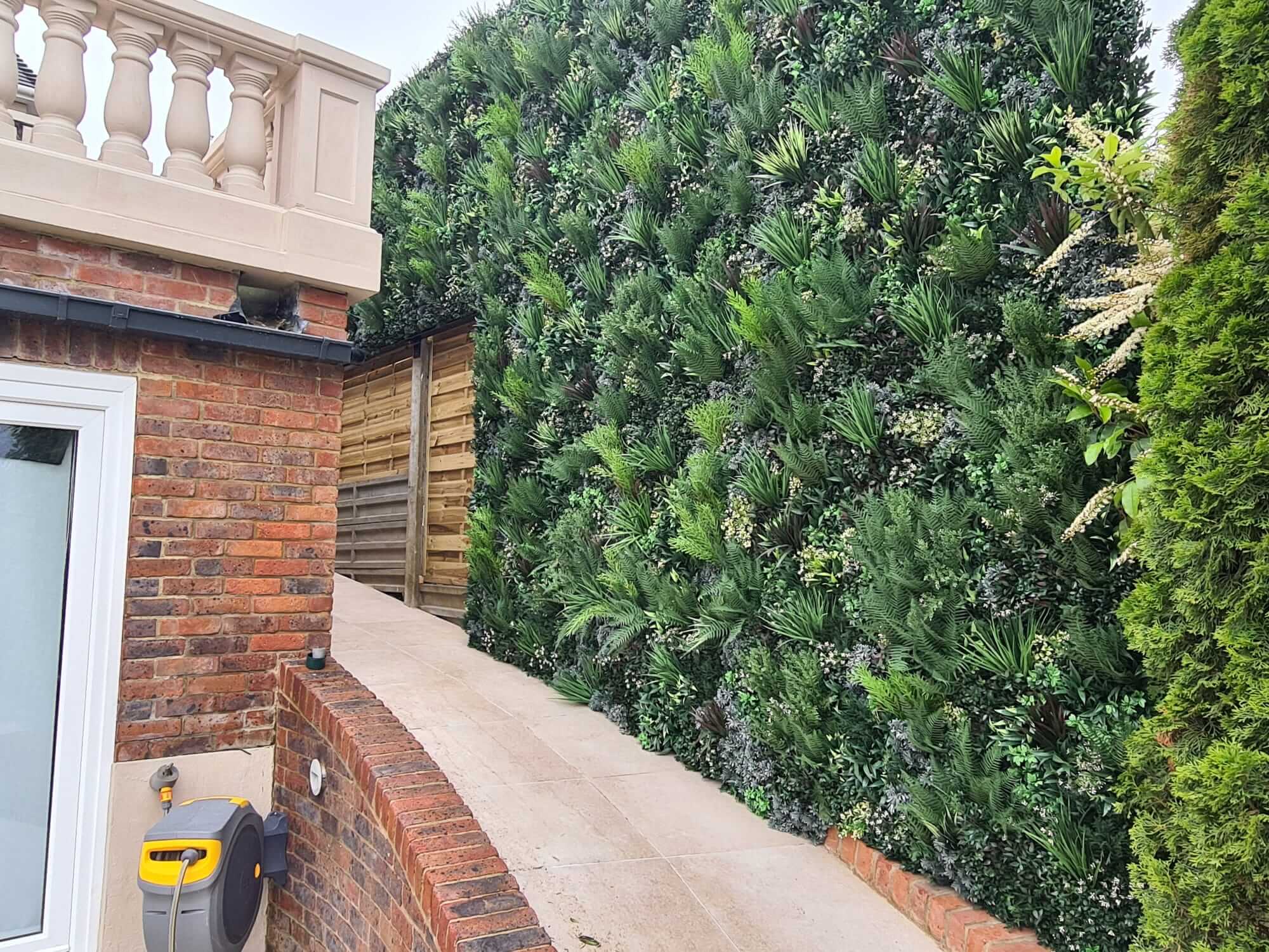 A narrow pathway bordered by a vibrant artificial green wall privacy screen on the right and a brick building with a window to the left, guiding you toward a wooden fence beneath a stone railing.