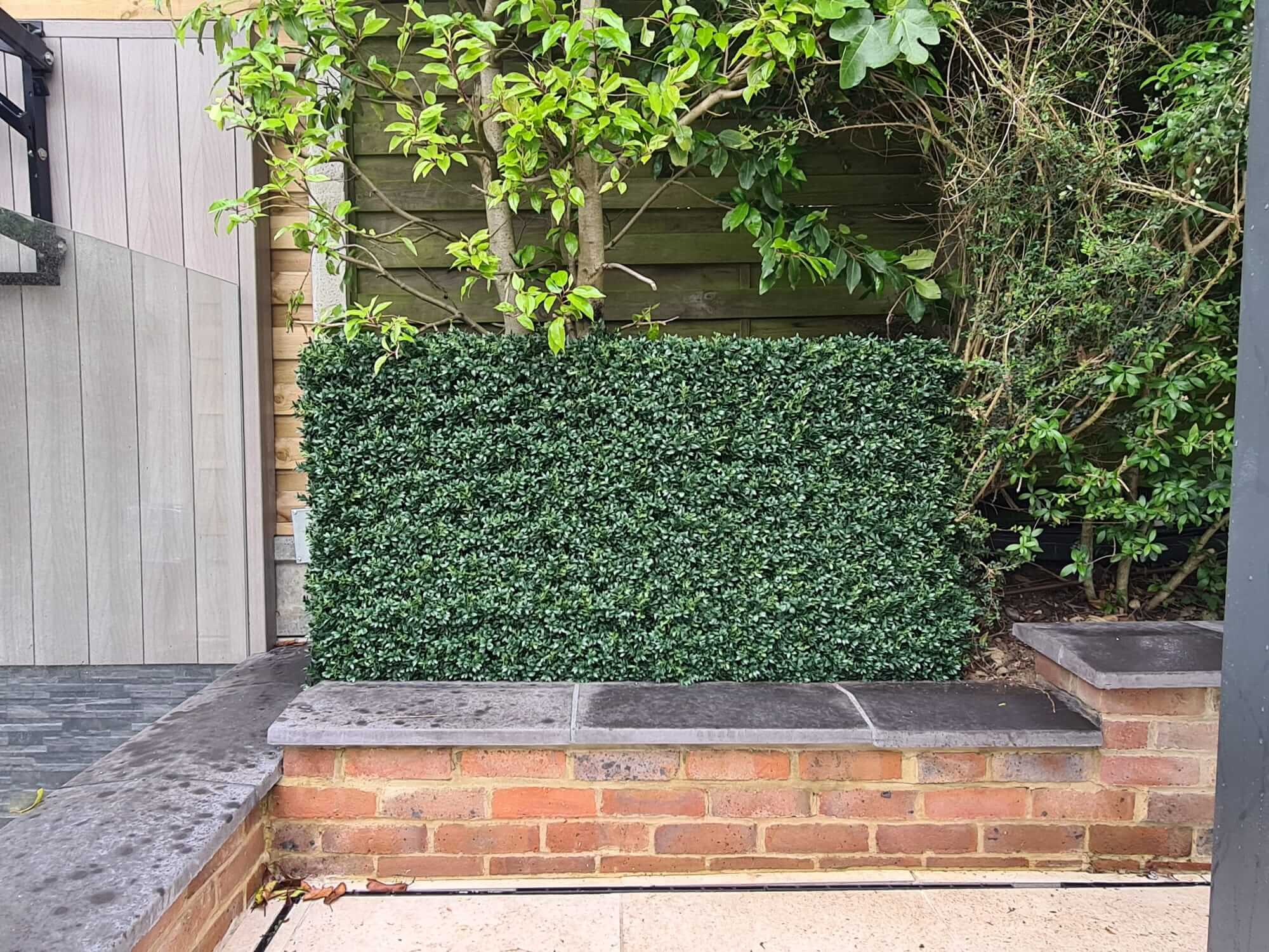 A bespoke artificial boxwood hedge installation sits on a raised brick planter, surrounded by a mix of green foliage. The ground is partly tiled and wet, suggesting recent rain. A wooden fence forms the backdrop, with contrasting modern wall paneling on one side.