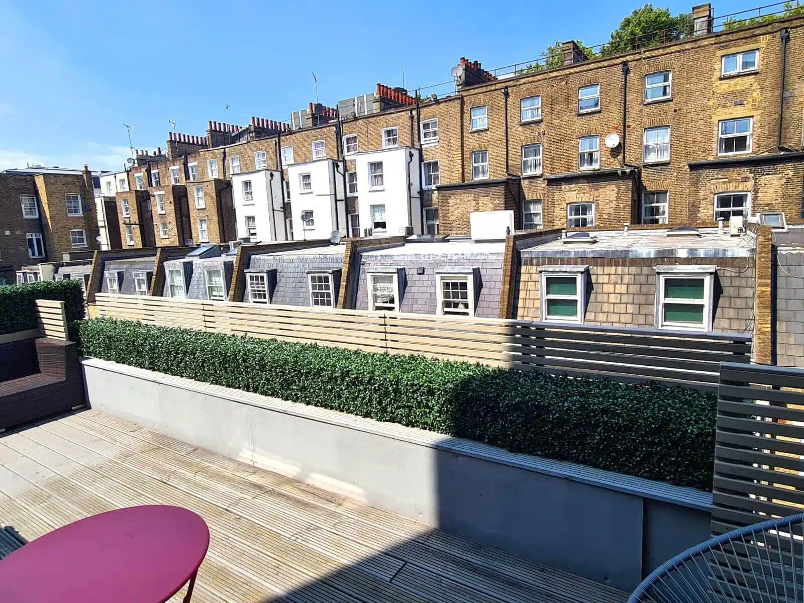 A rooftop terrace with red tables overlooks a row of brick buildings, their chimneys and skylights gleaming. The space features wooden decking and an artificial boxwood hedge installation along the edge. The sky is clear and blue.