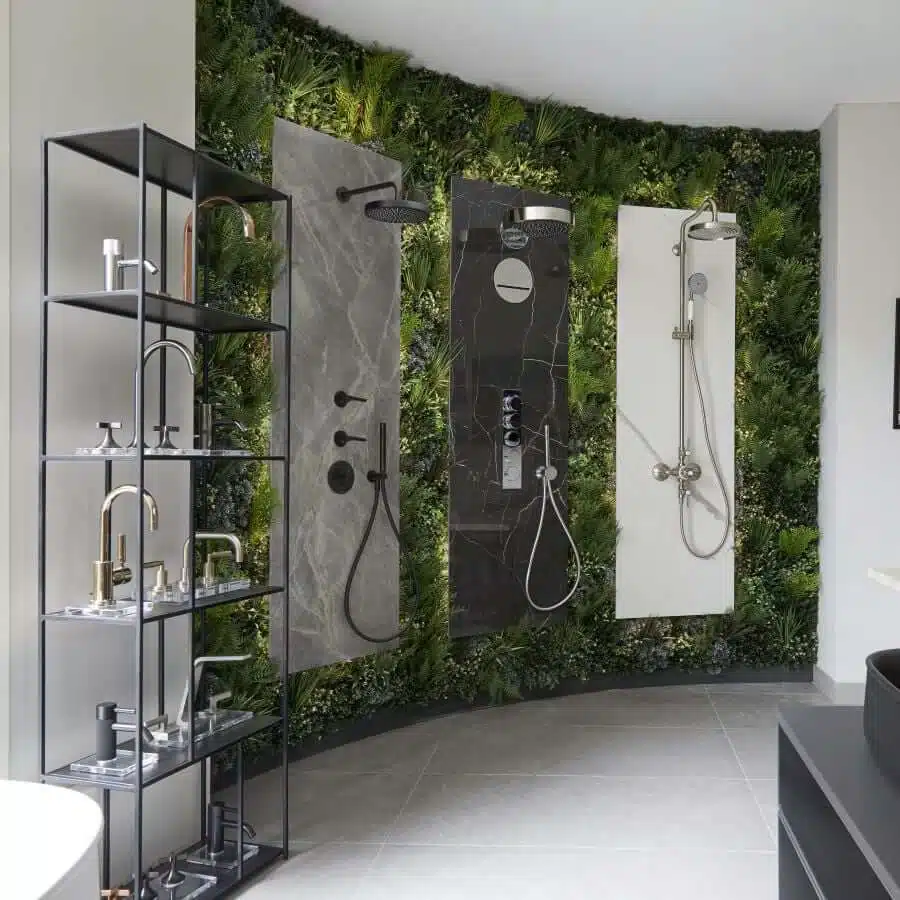 A display of three modern shower setups against a vibrant green wall in Windsors bathroom showroom. To the left, a black metal shelf holds various faucets. The light gray floor complements the scene, with a partial view of a sleek black sink on the right.