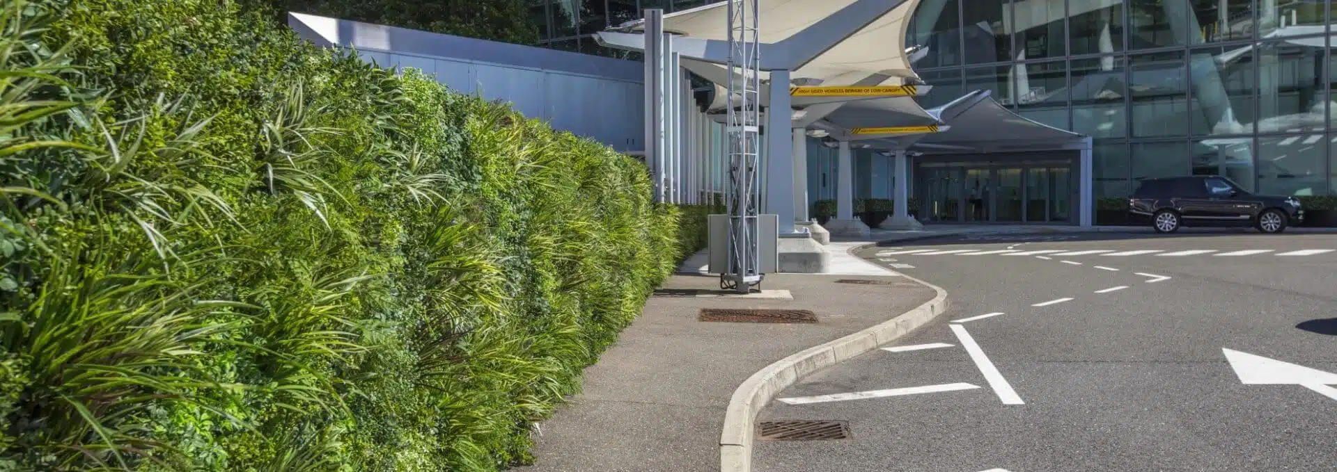A modern building entrance, reminiscent of Heathrow Airports Terminal 5 VIP Entrance, boasts a striking green wall on the left. A black car is parked on the right. Large glass windows define the structure, while a sleek canopy extends overhead. White lines neatly mark the road and path below.