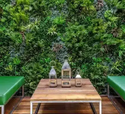 A vertical garden with lush green foliage transforms a London courtyard wall. In front, a wooden table holds three lanterns of varying sizes, evoking a serene atmosphere. Two green cushioned benches are placed on either side of the table, inviting you to relax and savor this urban oasis.