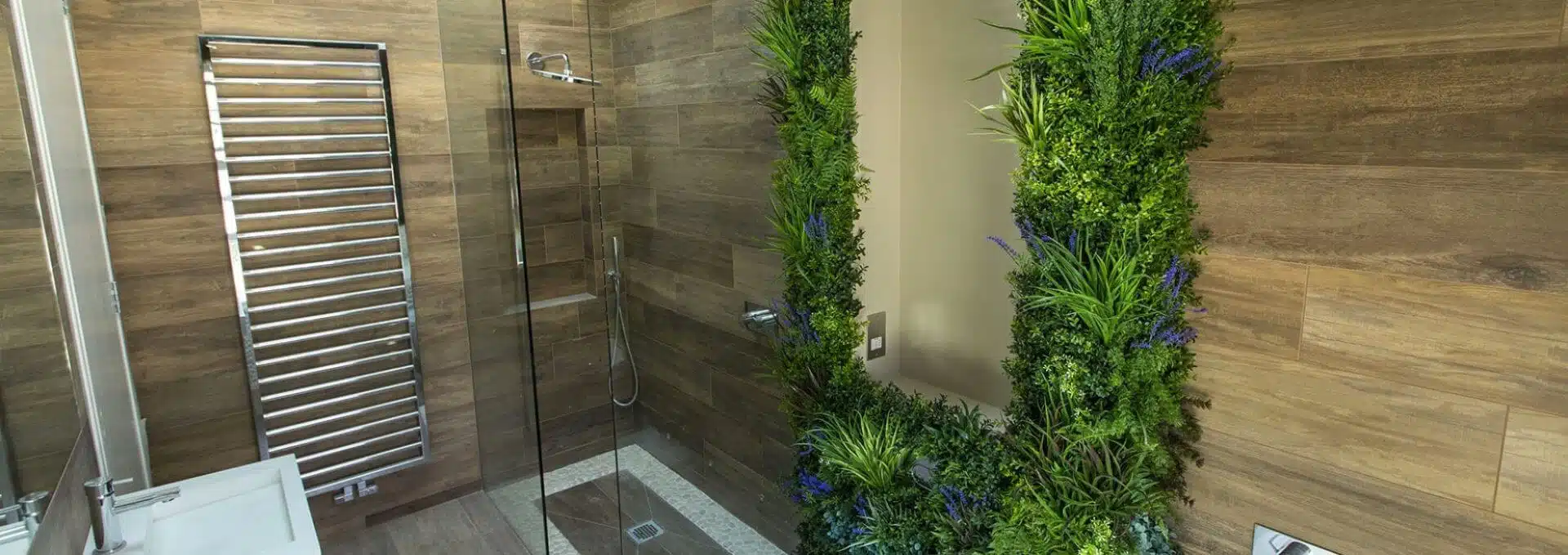 Modern bathroom featuring a wood-paneled design and a stunning bathroom green wall. It includes a glass shower enclosure, metal towel rack, and white sink. The lush vertical garden adds vibrant greenery with subtle hints of blue flowers.