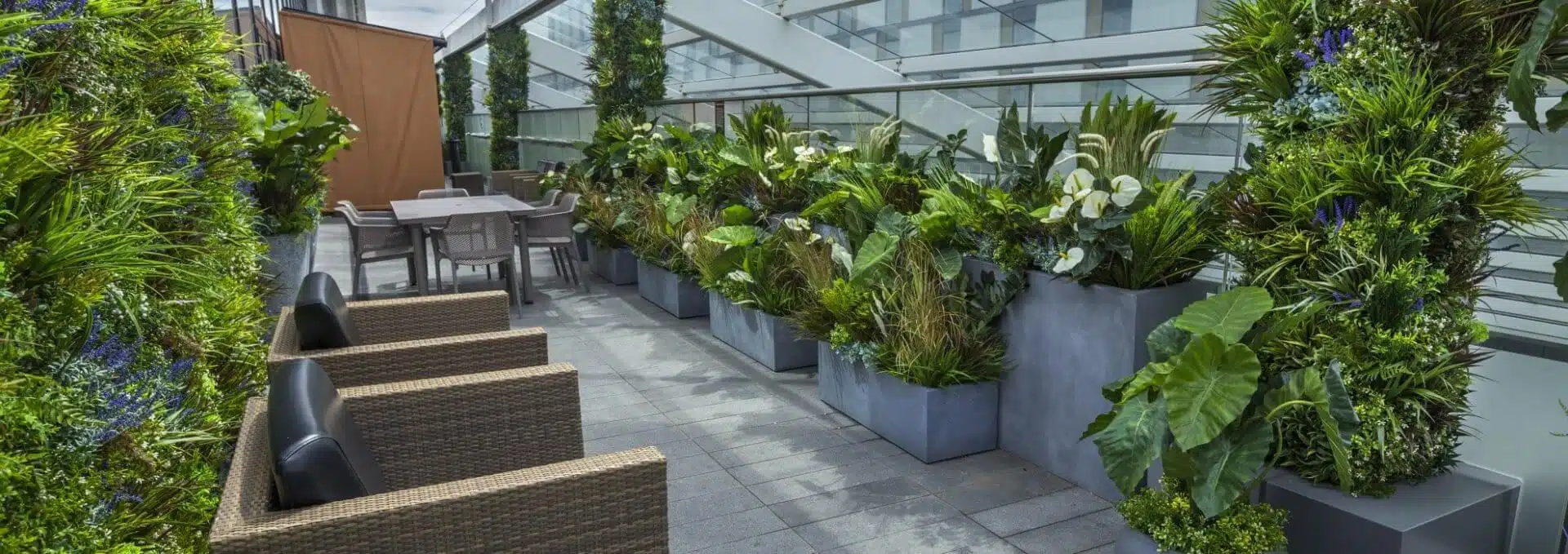 The rooftop garden overlooks London, with potted green plants lining a glass fence. Four wicker armchairs surround a table on the stone-tiled floor. In the background, a staircase and building facade contribute to this serene urban escape, reminiscent of the Casino Living Wall in Westfield Shopping Centre.
