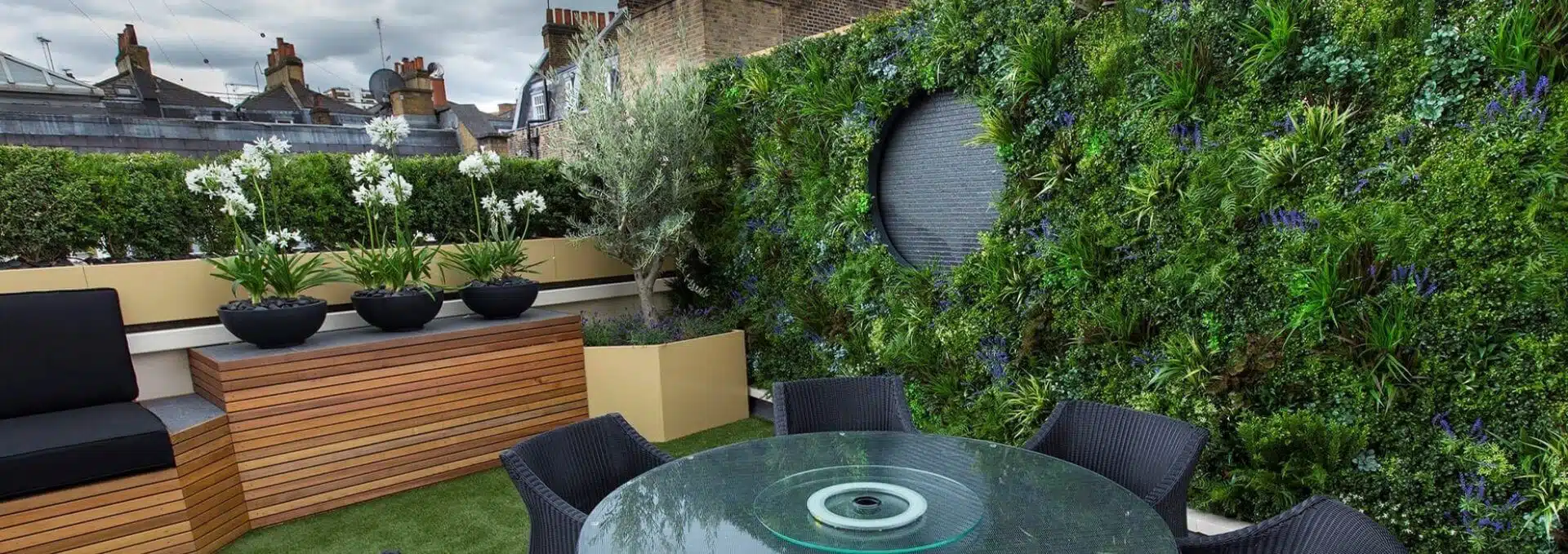 Terrace garden with a stunning vertical green wall showcasing diverse plants. A circular water feature graces the wall. On artificial turf, youll find a black cushioned bench, potted lilies, and a round glass-top table with black wicker chairs—all overlooking city rooftops.