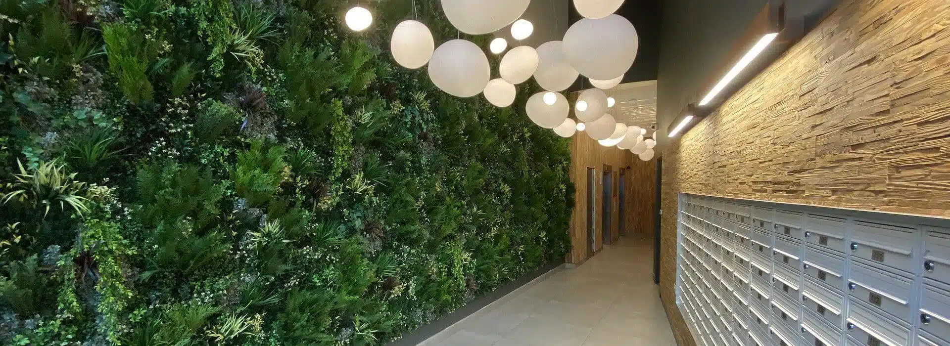 A hallway in Leeds, Yorkshire, boasts an artificial green wall on the left and mailboxes on the right. The ceiling showcases modern spherical pendant lights, while the floor is elegantly tiled.