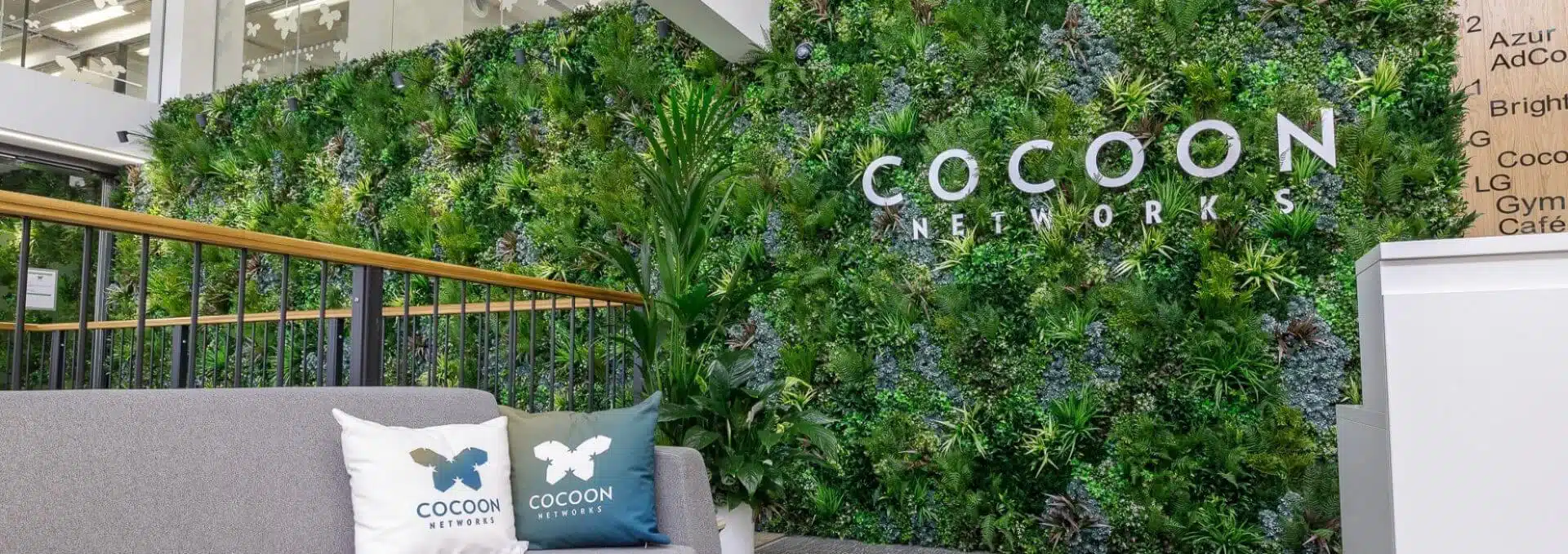 The office lobby boasts an artificial indoor green wall with the COCOON NETWORKS logo, adding a touch of nature in London. A gray sofa adorned with matching pillows sits in the foreground, while a staircase featuring a wooden railing stands on the left.