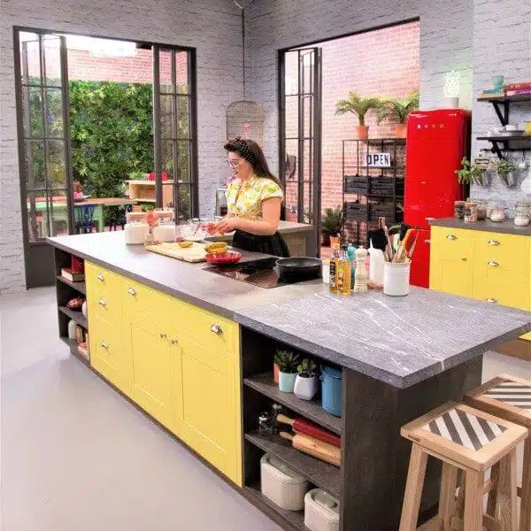 In a bright kitchen with a large island and yellow cabinets, a woman prepares food. The space, showcased on a popular TV show, features vertical green walls alongside the red fridge and potted plants, harmonizing with the cozy atmosphere created by the large windows.
