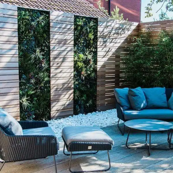 Modern outdoor patio with woven armchair, footrest, and round table set on a wooden deck in a green walled seating area in Manchester, UK. Vertical garden panels and bamboo plants bordered by horizontal wooden slats, with white stones adding greenery and texture.