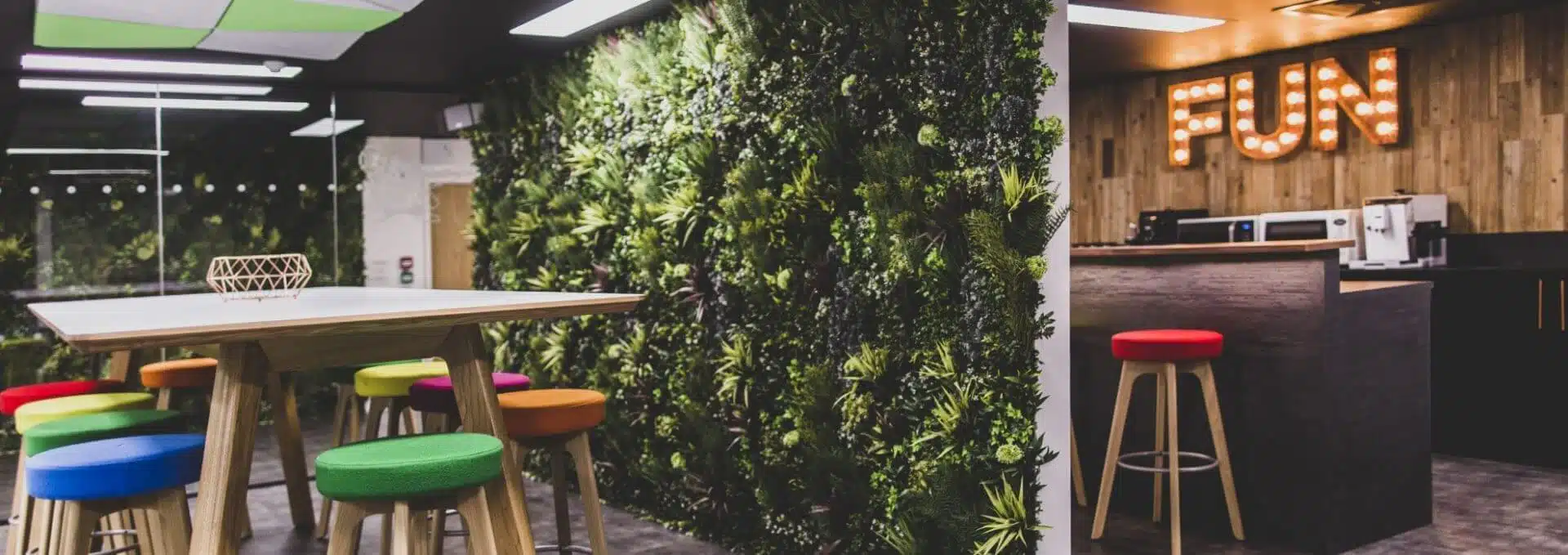 A modern indoor space in Guernsey boasts elegant green walls with a vertical plant display and a high table surrounded by colorful stools. In the background, a wooden counter and marquee letters spelling FUN adorned with bulbs create a lively and creative atmosphere.