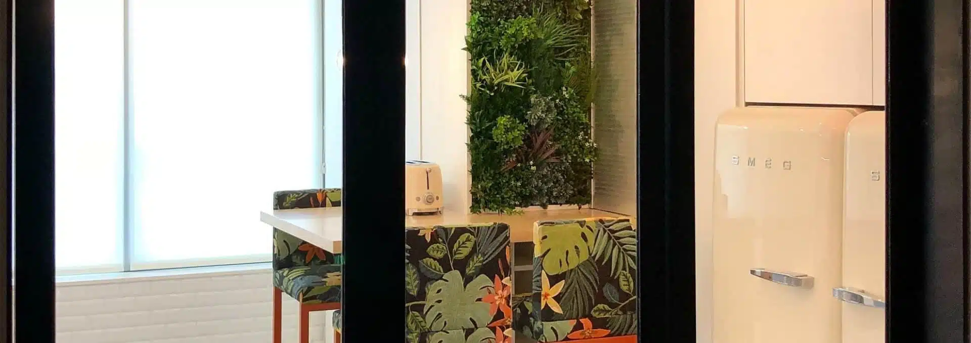 A modern kitchen viewed through glass doors, featuring a white retro-style fridge, a wooden table with floral-patterned chairs, and a green wall for privacy screening. A toaster sits on the table near the window, which emits soft natural light.